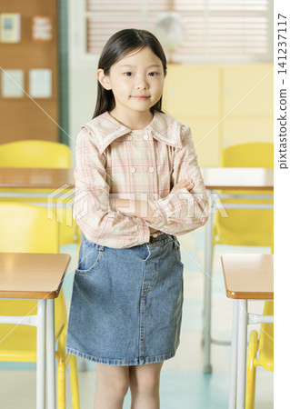 Smiling Asian schoolgirl posing with a backpack between classroom desks Smiling Asian schoolgirl posing with a backpack between classroom desks 141237117
