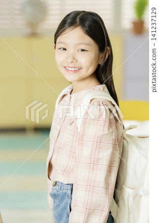 Smiling Asian schoolgirl posing with a backpack between classroom desks Smiling Asian schoolgirl posing with a backpack between classroom desks 141237129