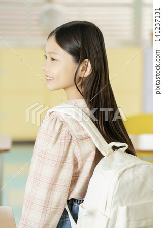 Elementary schoolgirl standing in a classroom with a white backpack Elementary schoolgirl standing in a classroom with a white backpack 141237131