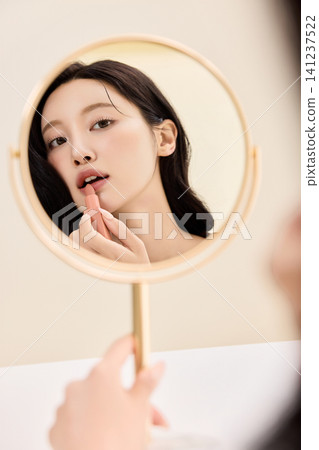 Young Asian woman applying makeup with a compact mirror Young Asian woman applying makeup with a compact mirror 141237522