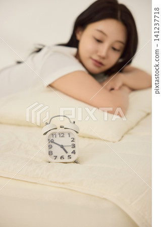 Asian woman lying on a bed and reaching toward an alarm clock 141237718