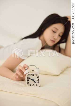 Woman resting on cream bedding beside a vintage alarm clock 141237720