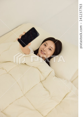 Young Asian woman using a smartphone while lying in bed Young Asian woman using a smartphone while lying in bed 141237763