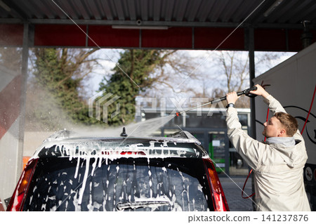 Young guy cleaning vehicle with pressure washer jet spray at manual car wash station. Young guy cleaning vehicle with pressure washer jet spray at manual car wash station. 141237876