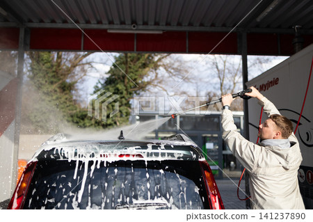 Young guy cleaning vehicle with pressure washer jet spray at manual car wash station. Young guy cleaning vehicle with pressure washer jet spray at manual car wash station. 141237890