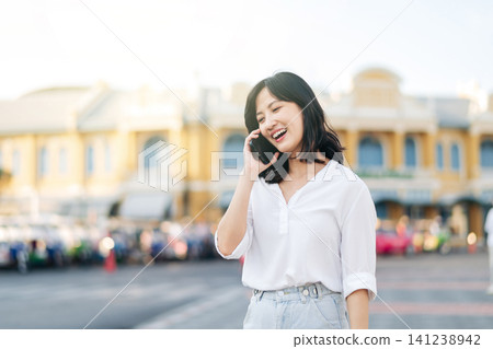 Portrait beautiful young asian woman calling on smart mobile phone standing on outdoor city street. Happy smiling lady talking outside on bright sunny summer day Portrait beautiful young asian woman calling on smart mobile phone standing on outdoor city street. Happy smiling lady talking outside on bright sunny summer day 141238942