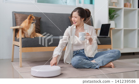 Smiling Asian woman operating robotic vacuum cleaner in living room with dog Smiling Asian woman operating robotic vacuum cleaner in living room with dog 141239586
