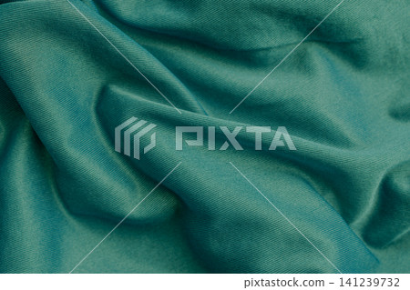 Horizontal close up shot of a smooth dark green or teal silky synthetic fabric texture draped in elegant soft wavy folds creating a luxury background 141239732