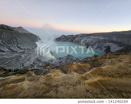 The stunning panoramic sunrise view of the turquoise acid crater lake at Mount Ijen volcano, featuring majestic rocky mountain ridges and thick sulfur smoke rising from the active vents in East Java. The stunning panoramic sunrise view of the turquoise acid crater lake at Mount Ijen volcano, featuring majestic rocky mountain ridges and thick sulfur smoke rising from the active vents in East Java. 141240284