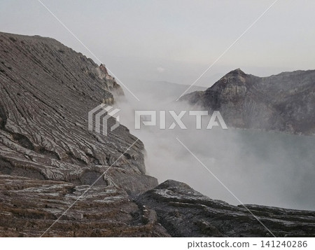 The stunning panoramic sunrise view of the turquoise acid crater lake at Mount Ijen volcano, featuring majestic rocky mountain ridges and thick sulfur smoke rising from the active vents in East Java. 141240286