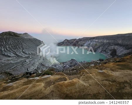 The stunning panoramic sunrise view of the turquoise acid crater lake at Mount Ijen volcano, featuring majestic rocky mountain ridges and thick sulfur smoke rising from the active vents in East Java. The stunning panoramic sunrise view of the turquoise acid crater lake at Mount Ijen volcano, featuring majestic rocky mountain ridges and thick sulfur smoke rising from the active vents in East Java. 141240357