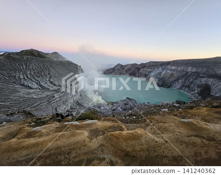 The stunning panoramic sunrise view of the turquoise acid crater lake at Mount Ijen volcano, featuring majestic rocky mountain ridges and thick sulfur smoke rising from the active vents in East Java. The stunning panoramic sunrise view of the turquoise acid crater lake at Mount Ijen volcano, featuring majestic rocky mountain ridges and thick sulfur smoke rising from the active vents in East Java. 141240362
