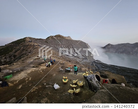 The stunning panoramic sunrise view of the turquoise acid crater lake at Mount Ijen volcano, featuring majestic rocky mountain ridges and thick sulfur smoke rising from the active vents in East Java. 141240372