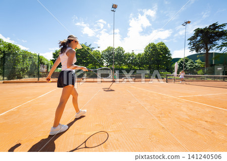Woman plays tennis on an outdoor clay court during a sunny day with trees and blue sky in the background Woman plays tennis on an outdoor clay court during a sunny day with trees and blue sky in the background 141240506