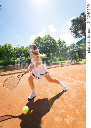 Person plays tennis on a clay court under a clear sky during the day with trees in the background and a bright sun above 141240509