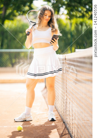 Young woman on tennis court checks phone while holding a racket and standing next to a net in bright light Young woman on tennis court checks phone while holding a racket and standing next to a net in bright light 141240556