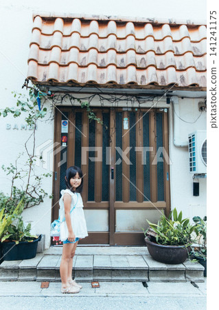 A young elementary school girl stands in front of a traditional Okinawan house with a red tiled roof. A young elementary school girl stands in front of a traditional Okinawan house with a red tiled roof. 141241175