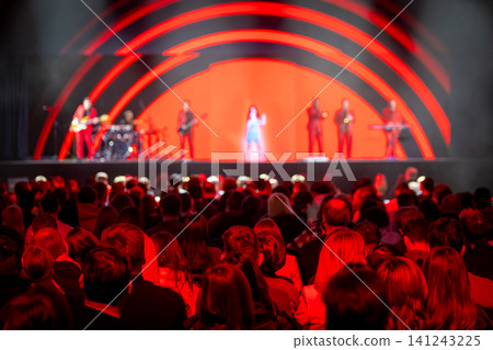 Live concert with band on stage under red lights, crowded audience enjoying performance Live concert with band on stage under red lights, crowded audience enjoying performance 141243225