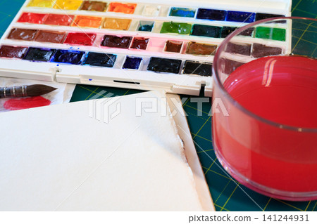 Watercolor paints palette with brush and blank paper sheet, glass jar with red tinted water on green cutting mat background 141244931