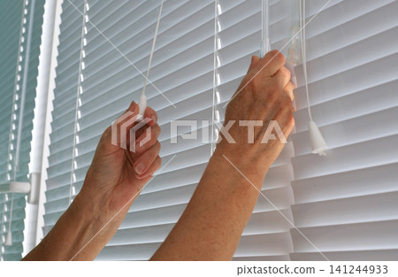 Hands adjusting white venetian window blinds using pull cords, person regulating sun light with horizontal plastic shutters Hands adjusting white venetian window blinds using pull cords, person regulating sun light with horizontal plastic shutters 141244933