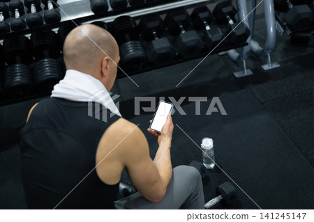 Athlete using smartphone while resting beside dumbbells in gym Athlete using smartphone while resting beside dumbbells in gym 141245147