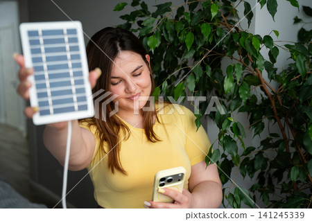 Young woman using smartphone while holding a portable solar panel indoors. Sustainable technology and off grid power innovation Young woman using smartphone while holding a portable solar panel indoors. Sustainable technology and off grid power innovation 141245339