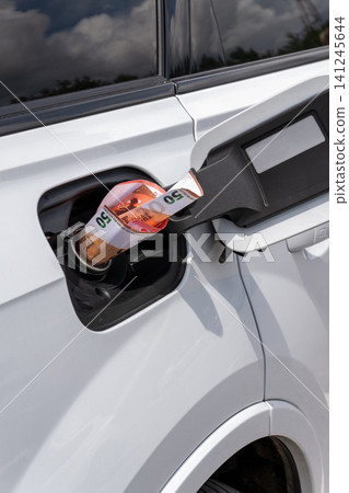 Rolled fifty Euro cash note inside vehicle fuel filler neck. Close-up view of white auto gas tank compartment with money symbolizing high petrol prices, inflation, and expensive transportation. Rolled fifty Euro cash note inside vehicle fuel filler neck. Close-up view of white auto gas tank compartment with money symbolizing high petrol prices, inflation, and expensive transportation. 141245644