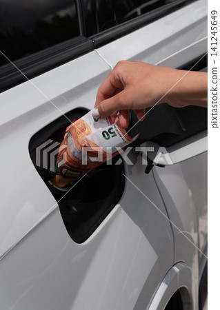 Female hand placing fifty Euro cash note inside vehicle fuel filler neck. Close-up view of white auto gas tank compartment with money symbolizing high petrol prices and rising transportation costs. Female hand placing fifty Euro cash note inside vehicle fuel filler neck. Close-up view of white auto gas tank compartment with money symbolizing high petrol prices and rising transportation costs. 141245649