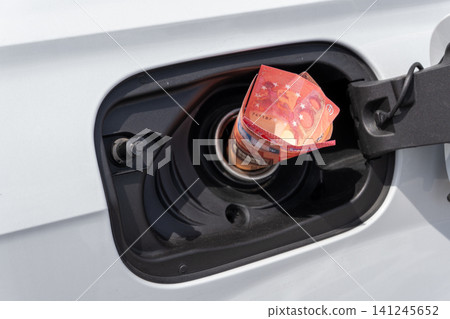 Rolled fifty Euro cash note inside vehicle fuel filler neck. Close-up view of white auto gas tank compartment with money symbolizing high petrol prices, inflation, and expensive transportation. Rolled fifty Euro cash note inside vehicle fuel filler neck. Close-up view of white auto gas tank compartment with money symbolizing high petrol prices, inflation, and expensive transportation. 141245652