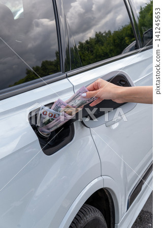 Female hand placing cash money inside vehicle fuel filler neck. Close-up view of white auto gas tank compartment filled with American dollar bills symbolizing high petrol prices and fuel cost. 141245653