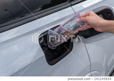 Female hand placing cash money inside vehicle fuel filler neck. Close-up view of white auto gas tank compartment filled with American dollar bills symbolizing high petrol prices and fuel cost. 141245654