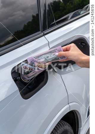 Female hand placing cash money inside vehicle fuel filler neck. Close-up view of white auto gas tank compartment filled with American dollar bills symbolizing high petrol prices and fuel cost. Female hand placing cash money inside vehicle fuel filler neck. Close-up view of white auto gas tank compartment filled with American dollar bills symbolizing high petrol prices and fuel cost. 141245656