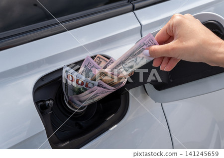 Female hand placing cash money inside vehicle fuel filler neck. Close-up view of white auto gas tank compartment filled with American dollar bills symbolizing high petrol prices and fuel cost. 141245659