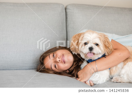 Teenage girl lying on grey couch and hugging her small fluffy dog 141245836