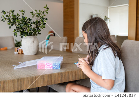Young girl sitting at wooden table and making jewelry with colorful beads Young girl sitting at wooden table and making jewelry with colorful beads 141245837