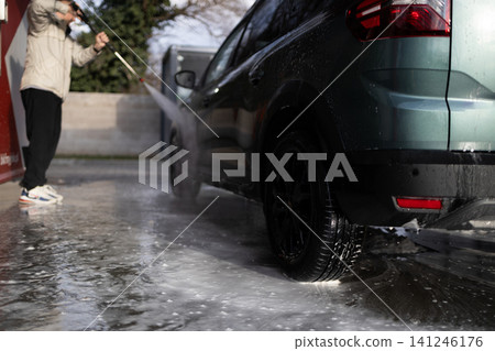 Young guy cleaning modern crossover vehicle with pressure washer jet at manual car wash. Young guy cleaning modern crossover vehicle with pressure washer jet at manual car wash. 141246176