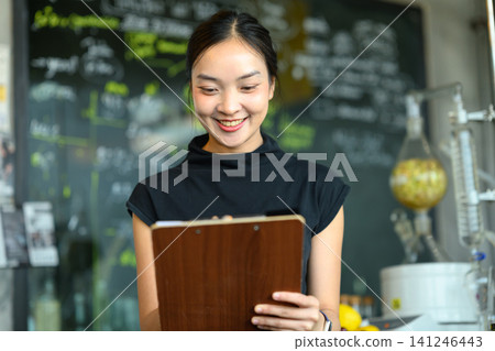 Young Asian Woman Smiling While Holding Clipboard in Cafe or Small Business Setting Young Asian Woman Smiling While Holding Clipboard in Cafe or Small Business Setting 141246443