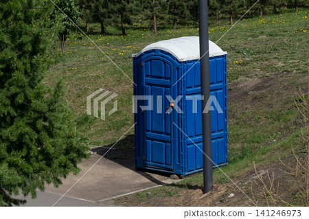 Bright Blue Portable Toilet Sitting in a Green Park Landscape Surrounded by Trees and Grass Under Clear Skies Bright Blue Portable Toilet Sitting in a Green Park Landscape Surrounded by Trees and Grass Under Clear Skies 141246973