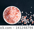 Coarse pink himalayan salt in a small white ceramic bowl on black slate background. High angle top view flat lay composition with sharp details and copy space. Perfect for cooking or wellness 141248794