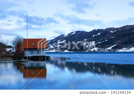 Oberstaufen, Germany, January 01 2018: Frozen calm lake with snowy mountains and reflected house 141249134