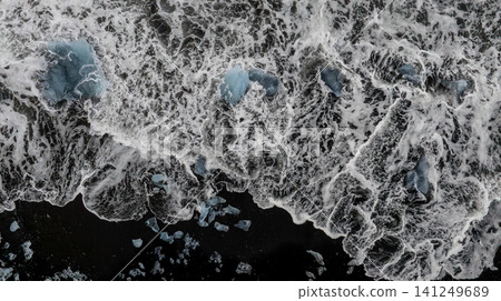 Aerial view of glacier ice fragments scattered across the black sands of Diamond Beach in Iceland, where powerful ocean waves create striking natural patterns and dramatic Arctic contrast. Aerial view of glacier ice fragments scattered across the black sands of Diamond Beach in Iceland, where powerful ocean waves create striking natural patterns and dramatic Arctic contrast. 141249689