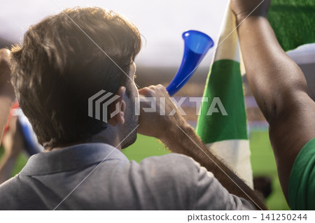 Close up of man in football stadium blowing air horn, live sports atmosphere concept. Excited fan creates loud crowd energy, team spirit and using vuvuzela Close up of man in football stadium blowing air horn, live sports atmosphere concept. Excited fan creates loud crowd energy, team spirit and using vuvuzela 141250244