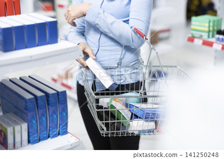 Closeup of woman placing medicine boxes into shopping basket while standing in pharmacy aisle, highlighting careful product selection, everyday healthcare decisions and responsible drugstore purchase. Closeup of woman placing medicine boxes into shopping basket while standing in pharmacy aisle, highlighting careful product selection, everyday healthcare decisions and responsible drugstore purchase. 141250248