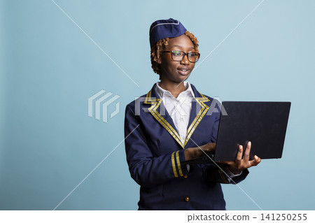 African american air hostess standing with laptop in hand. Female cabin crew member browses travel information and prepares for upcoming flight, showing professionalism in airline service and tourism. 141250255