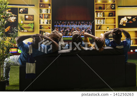 Soccer fans on couch putting hands on head after missed goal chance during TV match broadcast on screen. Rear view of football supporters showing worried reaction Soccer fans on couch putting hands on head after missed goal chance during TV match broadcast on screen. Rear view of football supporters showing worried reaction 141250263