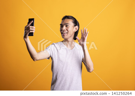 Cheerful Asian man holds smartphone and waves while smiling in front of yellow background. Friendly young adult enjoys video call, showing online communication, connection and positive lifestyle. 141250264