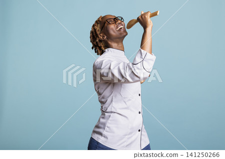 Cheerful female cook stands against blue background, playfully using wooden spoon as microphone. Joyful culinary expert singing happily with kitchen utensil, having fun performance in chef uniform. Cheerful female cook stands against blue background, playfully using wooden spoon as microphone. Joyful culinary expert singing happily with kitchen utensil, having fun performance in chef uniform. 141250266