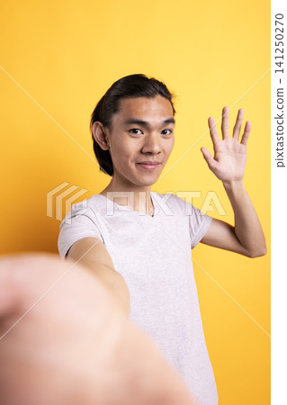 Smiling asian man waving at camera, filming personal vlog in studio. Cheerful male content creator greets online viewers with hello gesture, engaging warmly with social media audience. Smiling asian man waving at camera, filming personal vlog in studio. Cheerful male content creator greets online viewers with hello gesture, engaging warmly with social media audience. 141250270