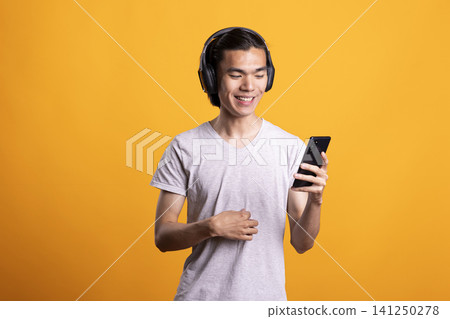 Smiling male model with headphones enjoys watching funny online content on smartphone. Cheerful man stands and reacts with amusement, streaming comedy clips and entertaining media on mobile device. Smiling male model with headphones enjoys watching funny online content on smartphone. Cheerful man stands and reacts with amusement, streaming comedy clips and entertaining media on mobile device. 141250278