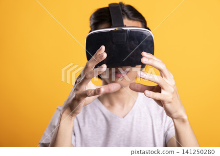 Asian man wears VR headset and reaches forward with hands, eating virtual burger. Person immersed in 3D digital world, enjoys playful augmented food interaction in studio with isolated background. Asian man wears VR headset and reaches forward with hands, eating virtual burger. Person immersed in 3D digital world, enjoys playful augmented food interaction in studio with isolated background. 141250280